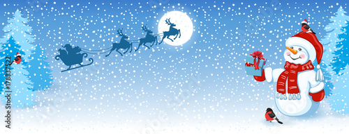  Snowman in Santa cap with gift box against winter forest background and Santa Claus in sleigh with reindeer team flying in the sky