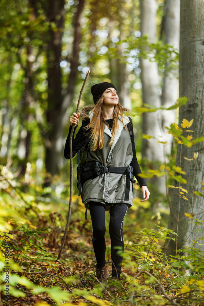 Cheerful brunette tourist girl wears black cap and backpacked have walk through forest holding stick, autumn tourism concept