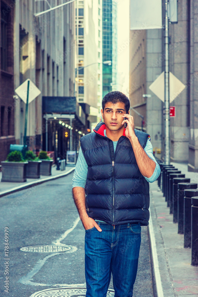 Modern City Life. East Indian American College Student wearing long sleeve  T shirt, blue down vest, jeans, walking on narrow street in New York,  talking on cell phone. Instagram filtered effect.. Stock