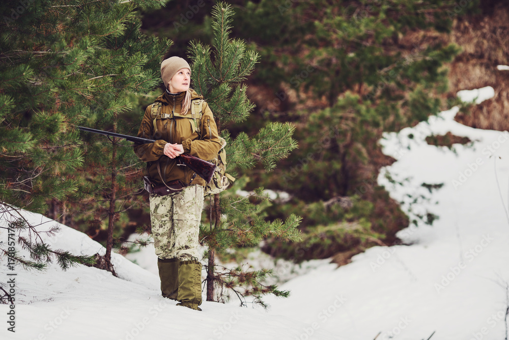 Female hunter in camouflage clothes ready to hunt, holding gun and walking in forest.