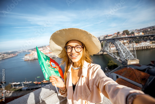 Young woman tourist making selfie photo with portuguese flag on the beautiful cityscape background during the morning light in Porto city, Portugal