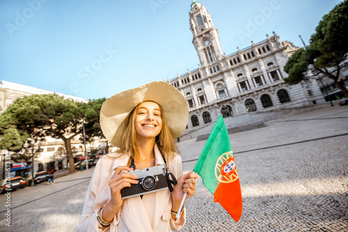 Portrait of a young woman tourist in sunhat standing with portuguese flag in front of the city hall building during the morning light in Porto, Portugal