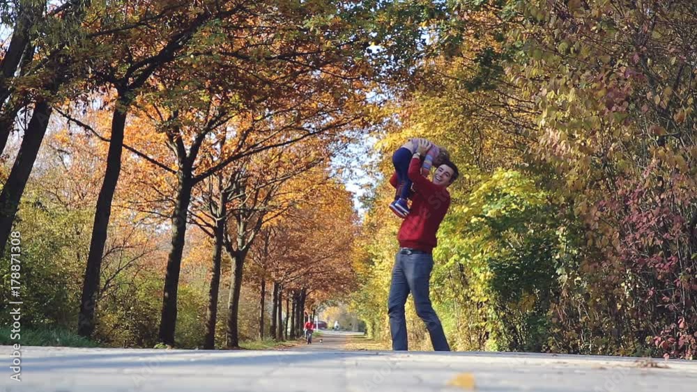 A father holds his daughter high up in the air and tosses her up and down. They stand beneath a colorful vault of autumn trees. A father holds his daughter high up in the air and tosses her up and down. They stand beneath a colorful vault of autumn trees.
