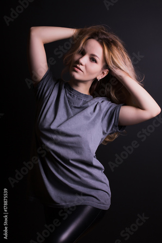 Fashionable young model with lush curly hair, wears grey shirt and leather leggins,  posing on a black background
