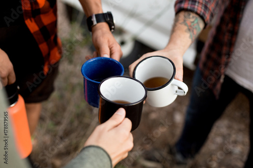 Three friends drink coffee or tea from hiking metal mugs or cups, hipsters with tattoos on adventure trip around europe, stop for rest or break to enjoy time together, hiking essensials