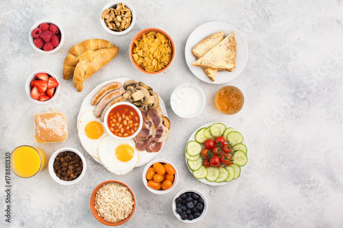 Healthy varied breakfast, fried eggs, sausages, bacon and mushrooms with selection of fruits and vegetables, breads and juice on the grey white table, top view, copy space for text, selective focus