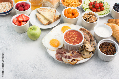 English breakfast made with fried eggs, sausages, bacon and mushrooms with selection of fruits and vegetables, breads and juice on the grey white table, copy space for text, selective focus