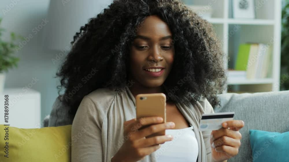 Curly African American young woman managing to buy using online shopping and credit card on smart phone. Home Curly African American young woman managing to buy using online shopping and credit card on smart phone. Home
