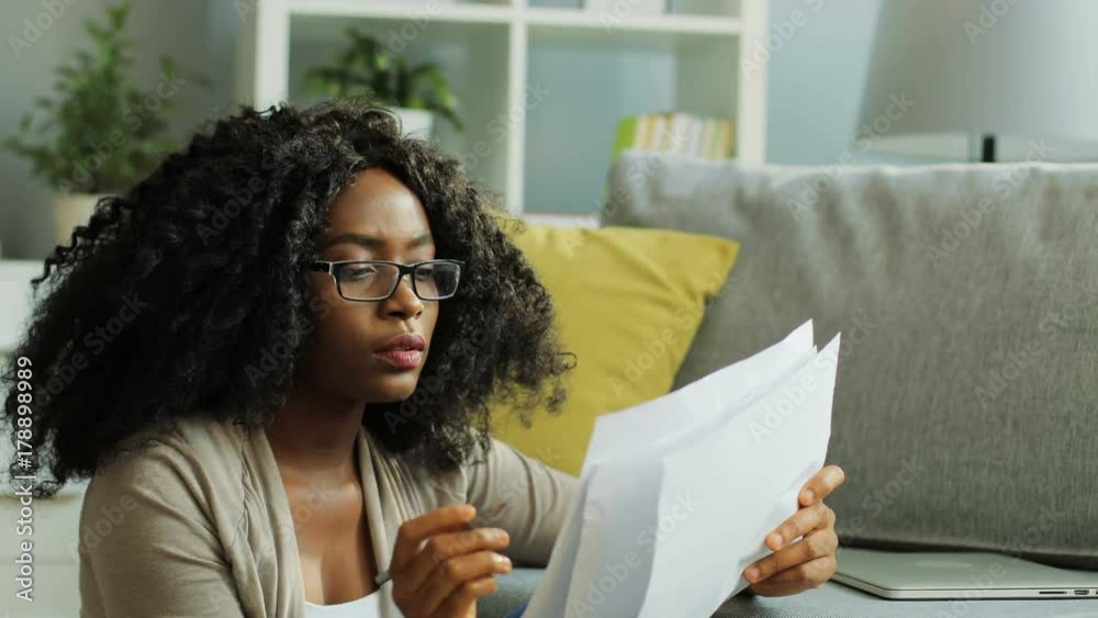 Portrait shot of beautiful curly young African American woman in glasses working with papper sheets, documents and pencil in the living room.