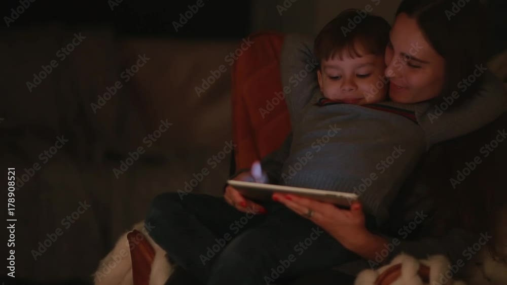 Beautiful woman mother sitting in a chair his home with son on his lap watching cartoons on the touch screen of the tablet. Beautiful woman mother sitting in a chair his home with son on his lap watching cartoons on the touch screen of the tablet.