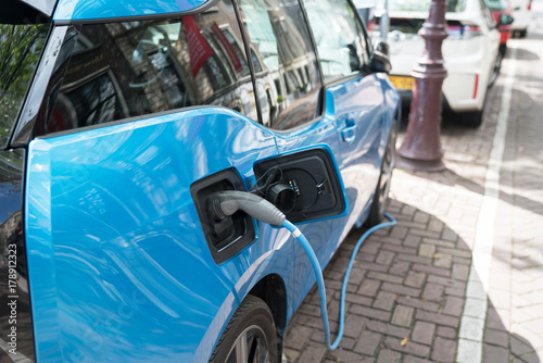 Blue electric car recharging on street with charge cable and plug leading to charge point.