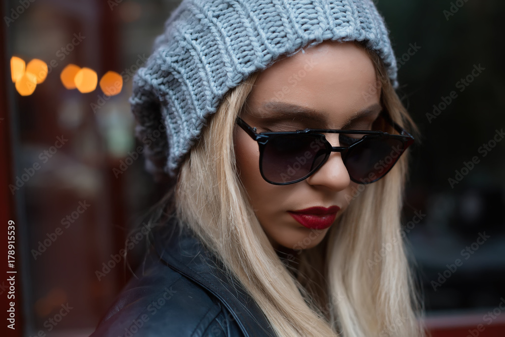 Close up female portrait of fashion model with long blonde curly hair wearing sunglasses sitting and posing outdoor. Jeans, sunglasses, hat, jacket.Young stylish woman Close up female portrait of fashion model with long blonde curly hair wearing sunglasses sitting and posing outdoor. Jeans, sunglasses, hat, jacket.Young stylish woman