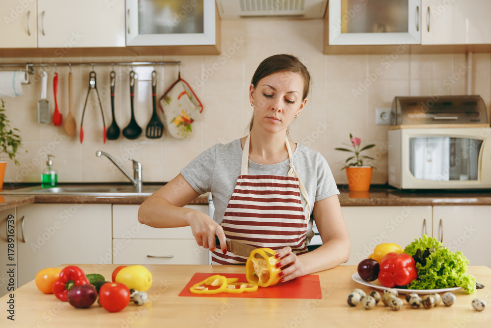 A young attractive woman in an apron cuts vegetables for salad with a knife in the kitchen. Dieting concept. Healthy lifestyle. Cooking at home. Prepare food.