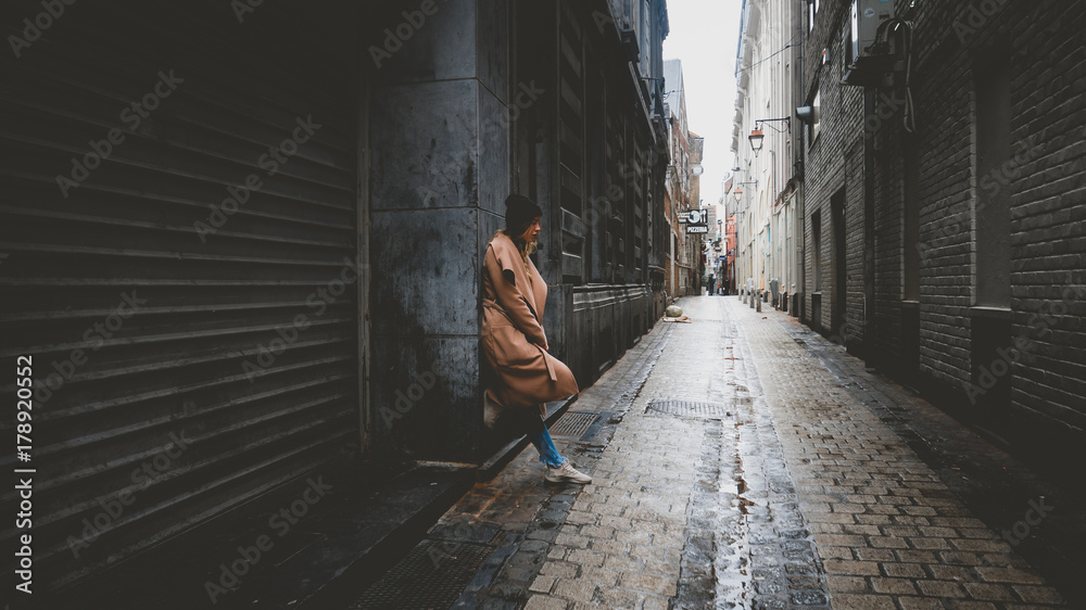 Lifestyle full portrait of young attractive fashionable Caucasian female in a coat standing in the narrow street in Europe. Horizontal orientation