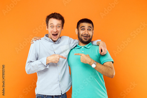 Two best friends hugging, pointing finger each other and looking at camera with shocked face photo
