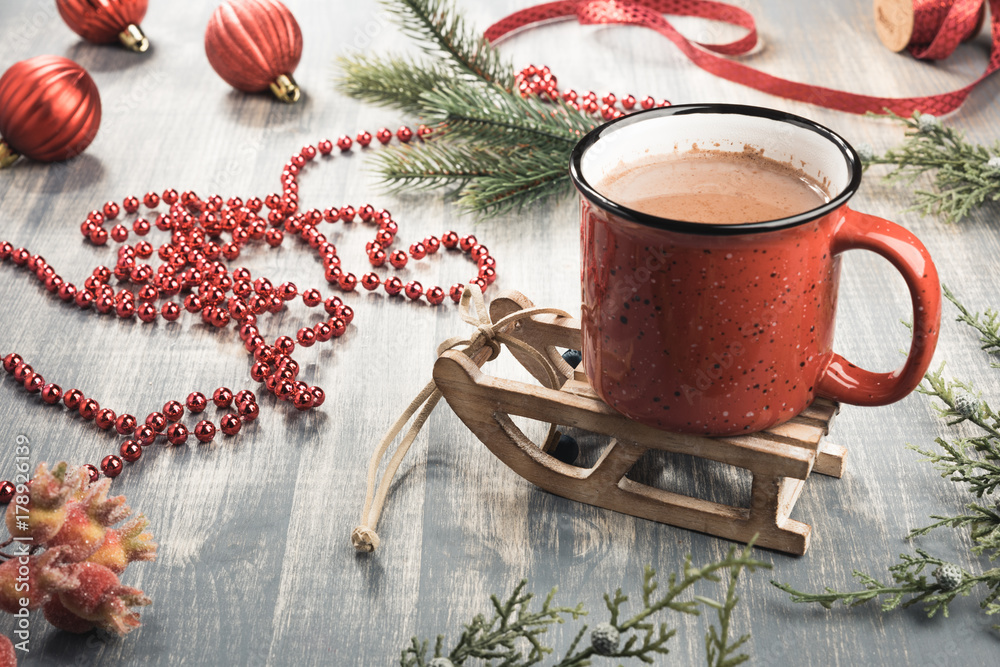 Cup of cocoa on grey wooden background. Red mug of hot chocolate on miniature toy sledge. Fir-tree branches, tape, Christmas balls Cup of cocoa on grey wooden background. Red mug of hot chocolate on miniature toy sledge. Fir-tree branches, tape, Christmas balls