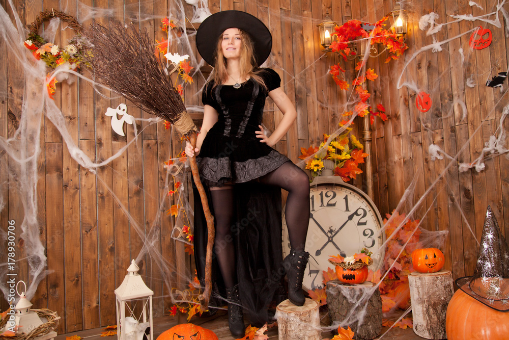 Fototapeta premium beautiful classic witch woman wearing black sorceress velvet dress and pointed hat, with orange evil halloween pumpkins and spiderweb 