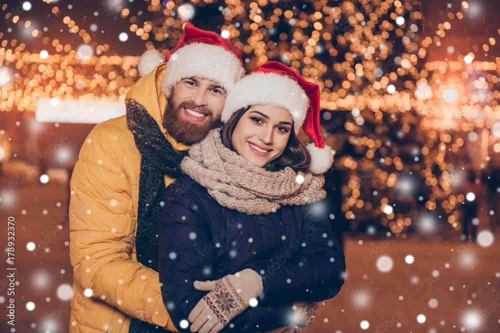 Portrait of happy cute young married couple hugging each other and smiling while walking at christmas eve outdoors, wearing santa`s hats, many lights, snowflakes and snow on the background Portrait of happy cute young married couple hugging each other and smiling while walking at christmas eve outdoors, wearing santa`s hats, many lights, snowflakes and snow on the background