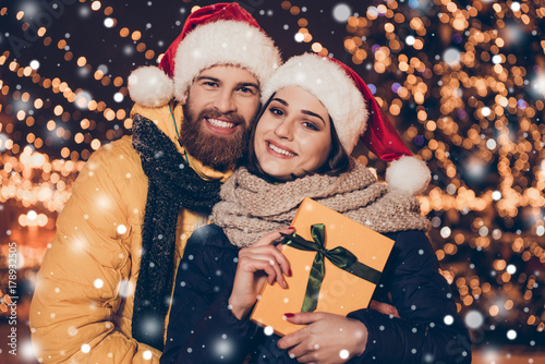Christmas miracle, joy, love, holy spirit! Young handsome red bearded man and his cute brunette lady, both smiling, happy, are looking at the camera, behind are many lights and snowfall of snowflakes