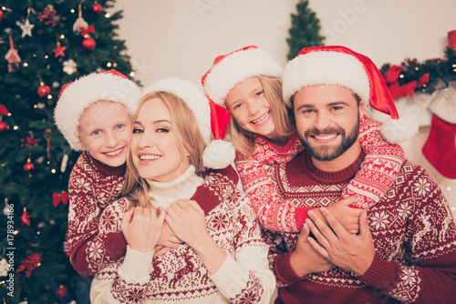 Portrait of four cheerful cuddling bonding relatives, at home, married couple, excited siblings ride mom and dad, in knitted cute traditional x mas costumes, pine firtree, happiness