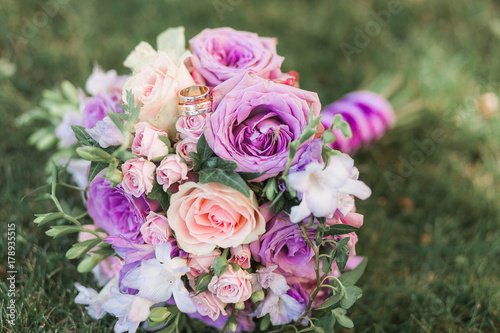 Beautiful wedding colorful bouquet for bride with golden rings on it lies on grass. Purple, white and peach flowers. Bridal accessories. Details for marriage and for married couple