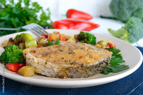 Steaks of fried spicy fish, served with a warm vegetable salad on a ceramic plate on a white wooden background.