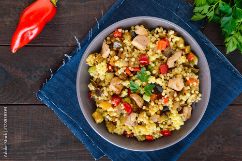 Light healthy dietary salad with couscous, vegetables (zucchini, eggplant, carrots, sweet peppers, onions), chicken pieces on a dark wooden background. Top view.