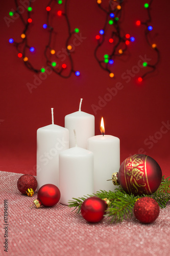 white candles with red christmas baubles on red background and led fairy lights