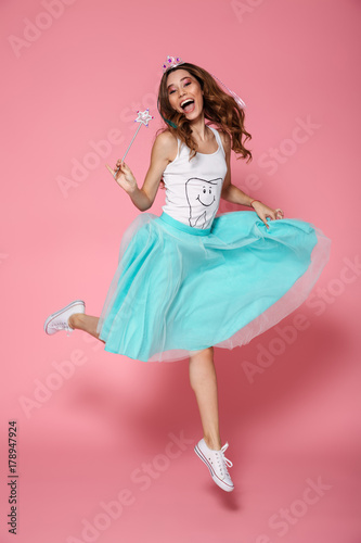Full length photo of cheerful princess, looking at camera, holding magic wand while jumping over pink background