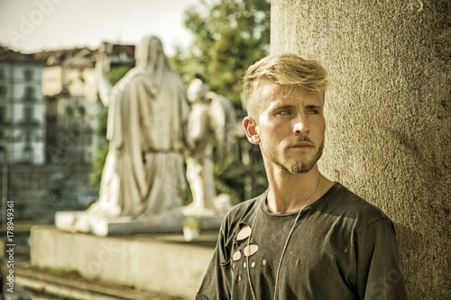 Handsome blond, blue eyed young man leaning against stone wall outdoor and looking to a side in Turin, Italy