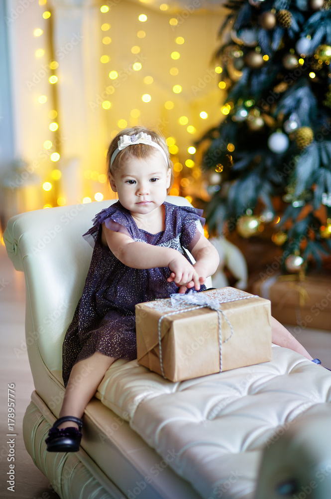 cute little girl in lilac dress sitting in a chair and opens box with present for background Christmas tree cute little girl in lilac dress sitting in a chair and opens box with present for background Christmas tree