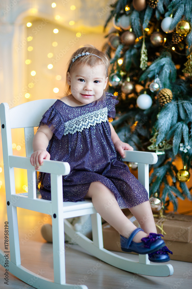 Very nice charming little girl blonde in lilac dress sitting on a child's stool and laughs loudly on the background of Christmas trees in bright interior of the house Very nice charming little girl blonde in lilac dress sitting on a child's stool and laughs loudly on the background of Christmas trees in bright interior of the house