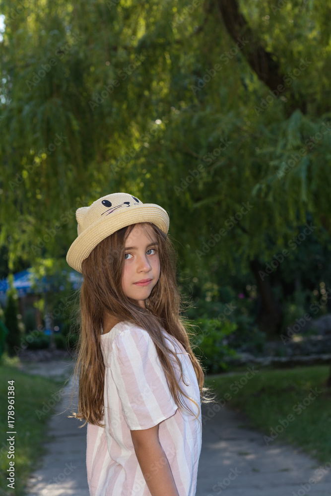 Cute little girl in a funny hat posing in the city park on a sunny summer day