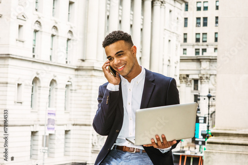 African American Businessman working in New York, wearing black blazer, white shirt, sitting by vintage office building, working on laptop computer, talking on cell phone. Instagram filtered effect..