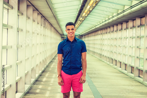 Summer in School. Wearing short sleeve shirt, red shorts, African American college student standing inside indoor walkway with glass walls, ceiling, wooden floor on campus in New York, smiling. . © Alexander Image