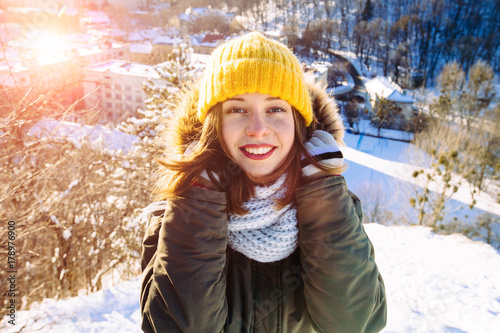 Young sporty girl in yellow knitting cap and scarf walking in the winter park on the background of snowy trees and city panorama. Female on the top of snowy hill in sunny day. photo