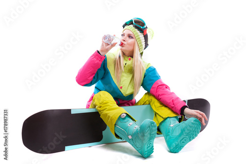 attractive young woman in ski suit and ski glasses sit on snowboard, drink water