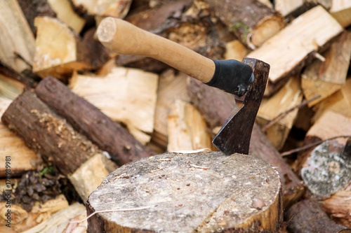 An axe on a wood, tree log. An axe stuck in a log in front of a pile of wood, ready for chopping and winter. Hardwood, wood industry. Heating season, winter season. Renewable resource of energy.