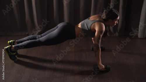 Athletic woman wearing sportswear is warming-up behind the scene. She is doing pushups with dumbbells indoor. photo