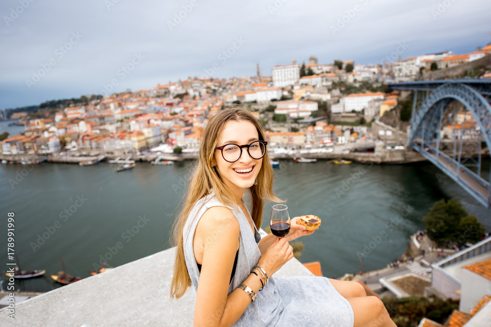 Portrait of a young woman enjoying traditional portuguese dessert called pastel de Nata, sitting on the terrace with beautiful cityscape view on Porto, Portugal