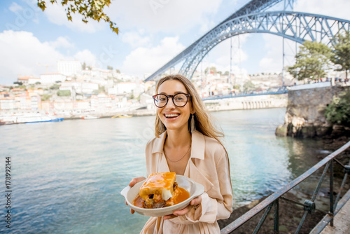 Portrait of a young woman with traditional portuguese sandwich with meat called francesinha on the landscape background with bridge in Porto city, Portugal
