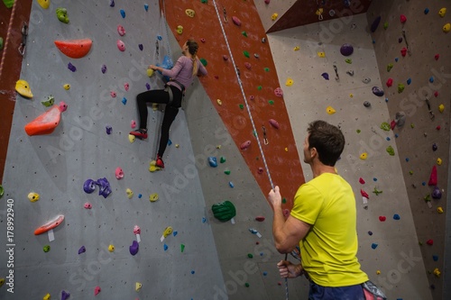Man holding rope looking at athlete climbing wall in fitness