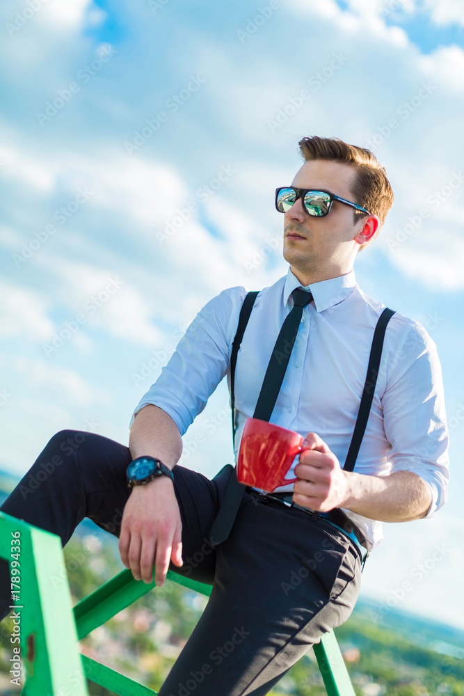 Fototapeta premium Attractive young busunessman in white shirt, tie, braces and sunglasses with red cup