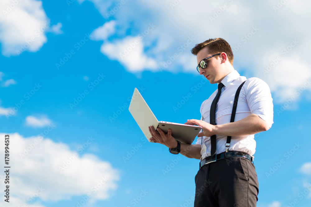 Attractive serious busunessman in white shirt, tie, braces and sunglasses on the roof with laptop Attractive serious busunessman in white shirt, tie, braces and sunglasses on the roof with laptop