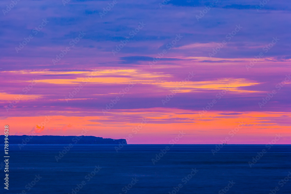 Fototapeta premium A thin coastline in the distance, in the foreground the blue sea, on the horizon the silhouette of the ship, a bright orange sky of a pink color. Colorful purple sunset at Bali, Indonesia.