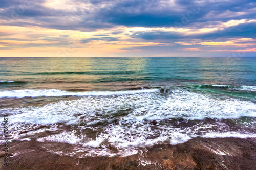 Foamy waves on a sunset background in cloudy weather with thunderclouds. Coastline of Tanah Lot, Beraban, Bali, Indonesia.