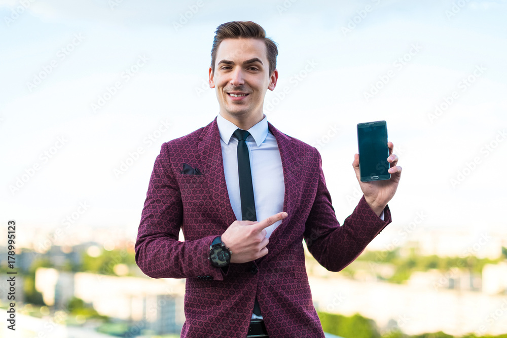 Young serious businessman in red suit and shirt with tie stand on the roof and show empty phone Young serious businessman in red suit and shirt with tie stand on the roof and show empty phone