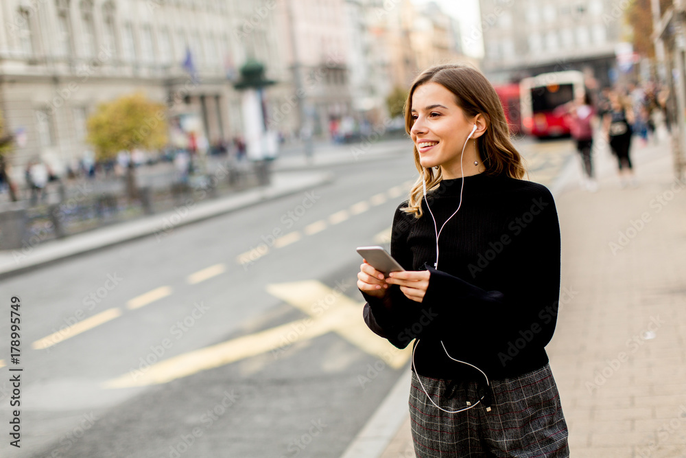 Fototapeta premium Young brunette woman using mobile while standing on street and waiting for a bus or taxi