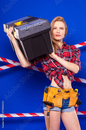 Young slim builder girl in red chechered shirt, builder belt, jean shorts and snickers hold toolbox photo