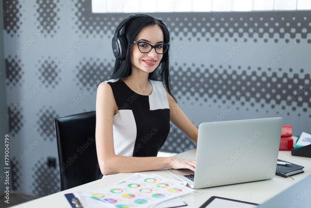 Beautiful young businesswoman in black dress, headphones and glasses sit at the table and work on laptop Beautiful young businesswoman in black dress, headphones and glasses sit at the table and work on laptop